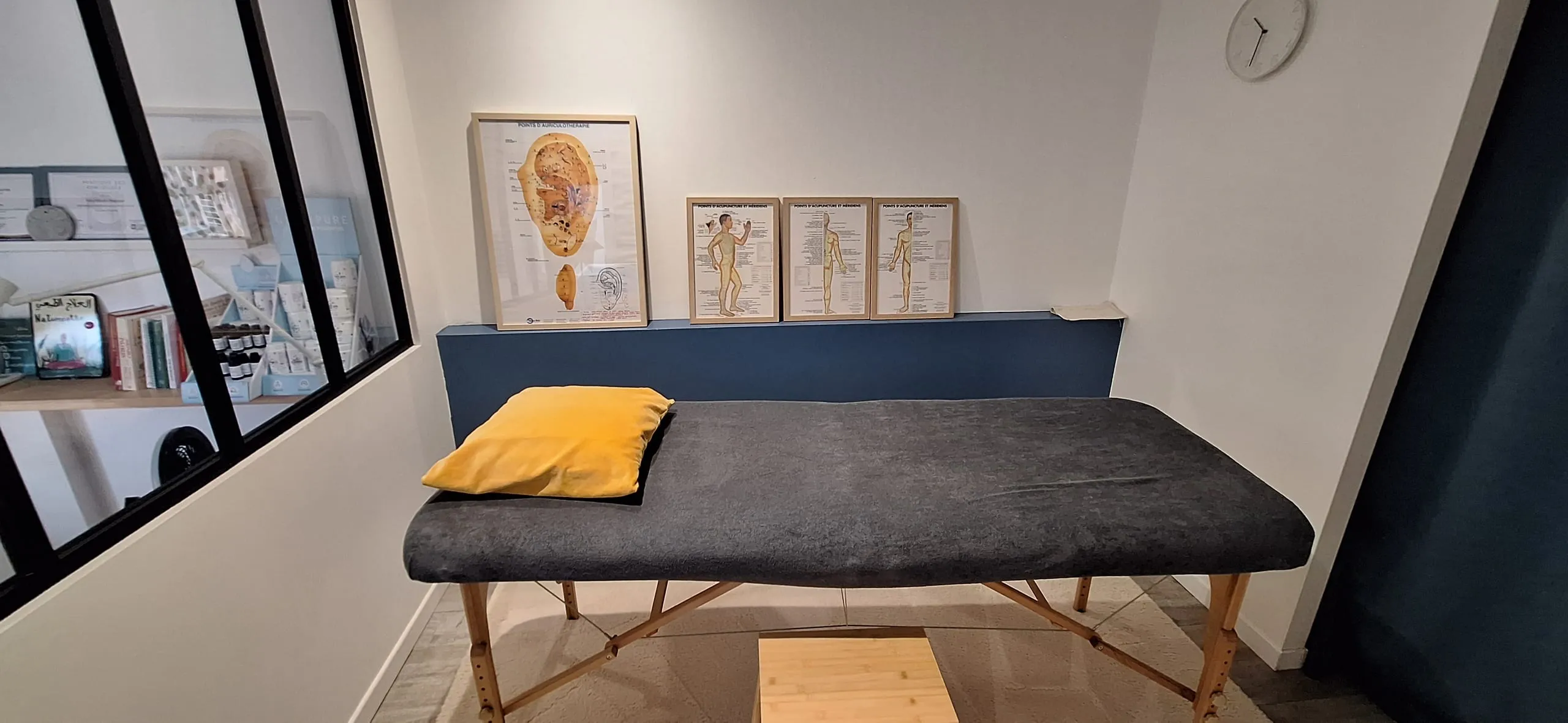 Salle de soin lumineuse avec table de massage, planches d’auriculothérapie et d’acupuncture sur un mur bleu et verrière.